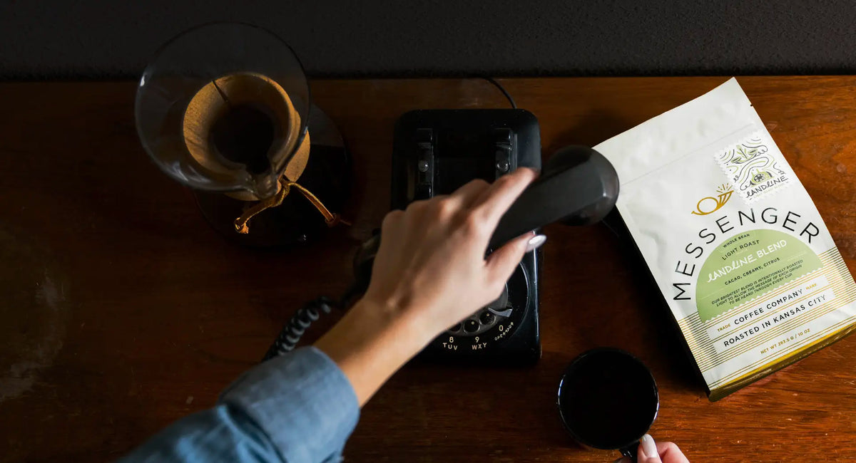 Person holding a phone receiver next to a bag of Messenger Coffee and a cup on a wooden table