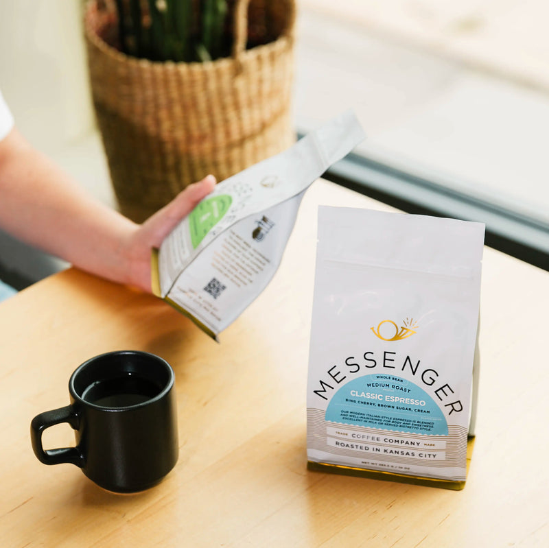 A person holding a bag of messenger coffee over a table surface. A bag of espresso and coffee cup in the foreground.