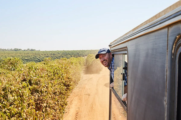 Man wearing cap leaning out of vehicle window on a dusty rural road with greenery on the side