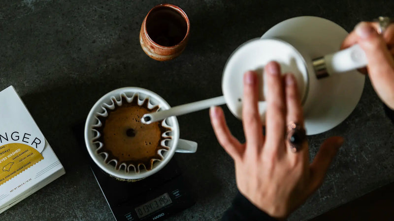 Person making coffee using a pour over method on a dark surface with a box of coffee and small container.