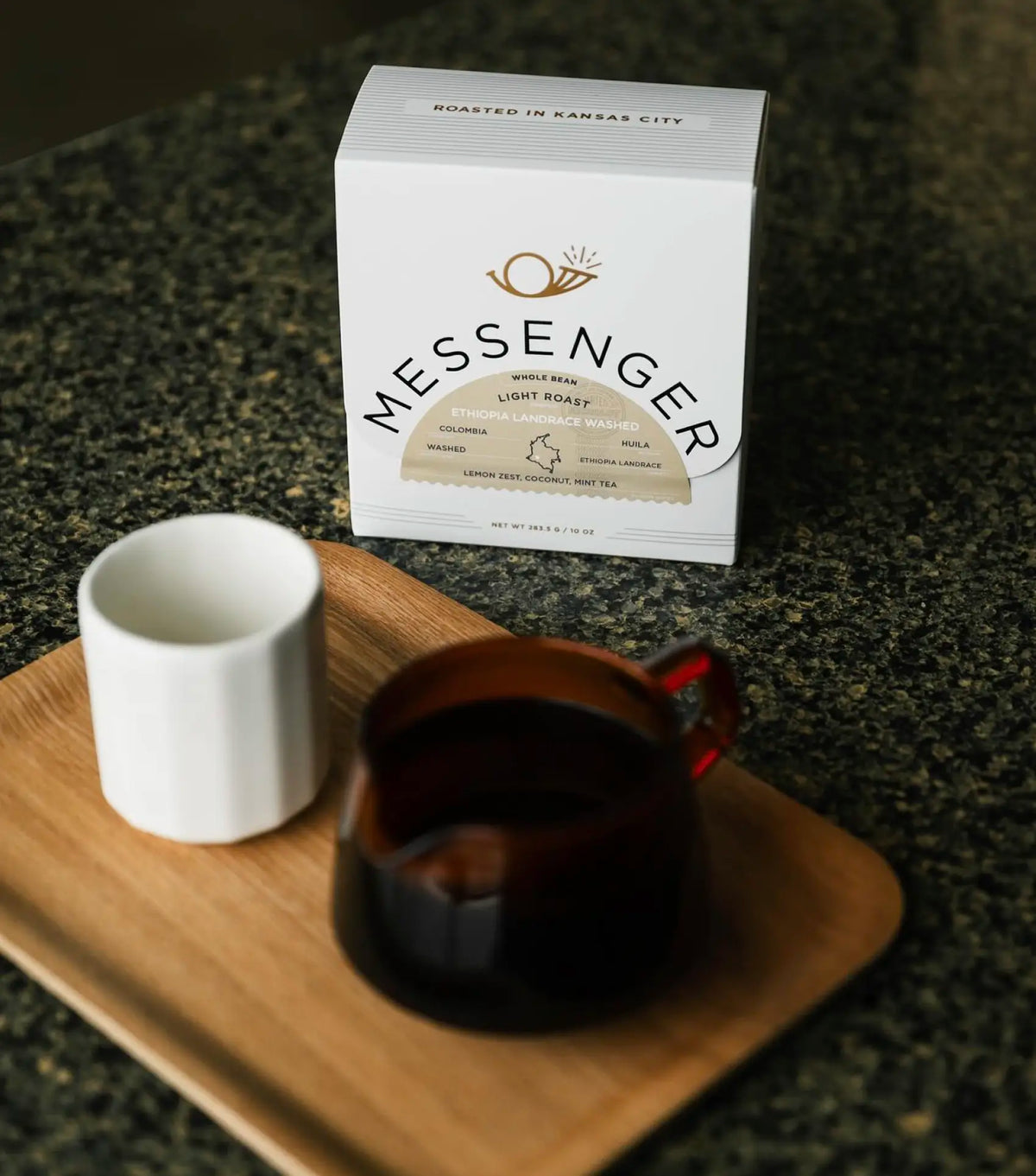 Box of Messenger light roast coffee with a dark glass cup and white ceramic mug on a wooden tray on a granite counter