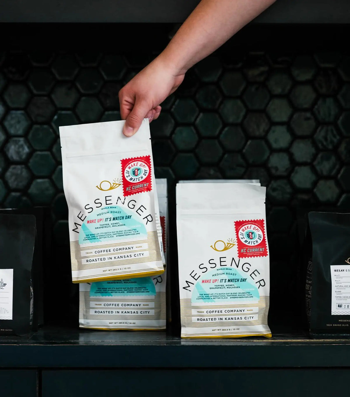 Hand placing a white bag of Messenger coffee on a shelf with other coffee bags in a dark tiled background