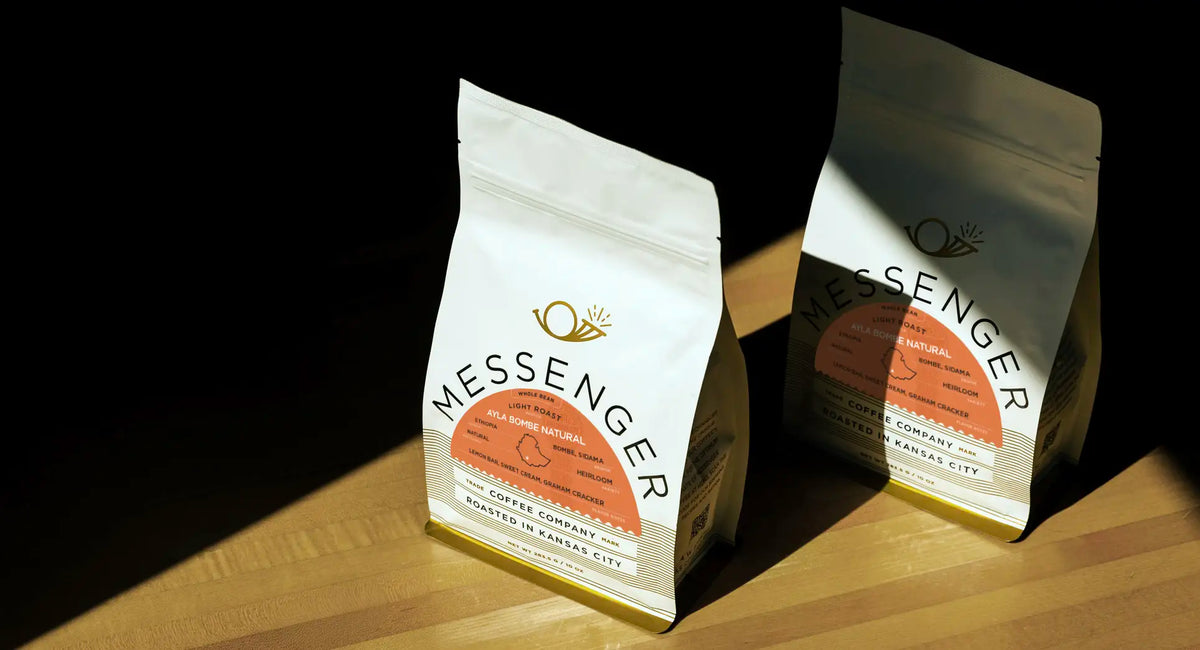 Two bags of Ayla Bombe Natural coffee with Messenger branding on a wooden surface and dramatic diagonal shadows.